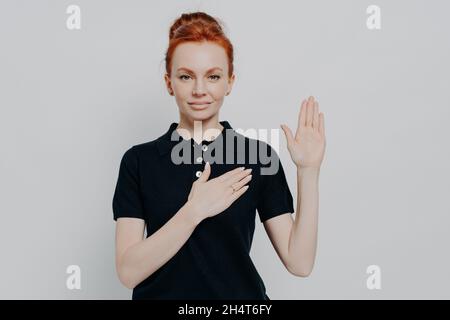 tudio shot of serious red haired female making promise, swearing with hand on chest in studio Stock Photo