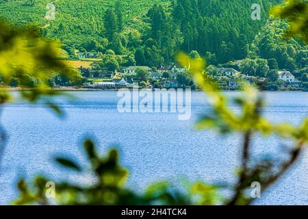 Loch Ness of Scotland in summer. View through branches of the lake with boat. Houses and trees in the background. Mountain slopes with different types Stock Photo