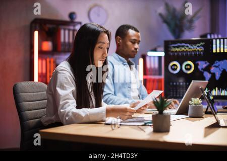 Asian woman and african american man sitting together at modern office and working on tablet and laptop. Competent economists analyzing charts on trade market during evening time. Stock Photo