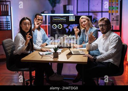 Four multi racial office workers showing okay sign and smiling on camera during business meeting. Coworkers using charts, graphs and technology while discussing common project. Stock Photo