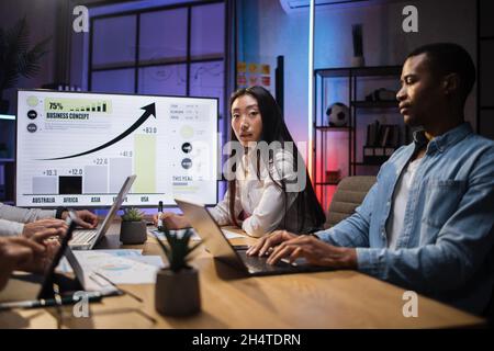 Four multiracial financial experts sitting at office with various gadgets and analysing financial growth in worldwide economy. Focus on Asian young lady posing on camera Stock Photo