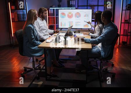 Woman presenting financial report of worldwide market on big monitor during meeting with international partners. Diverse colleagues sitting at desk and appealing to her speech. Stock Photo