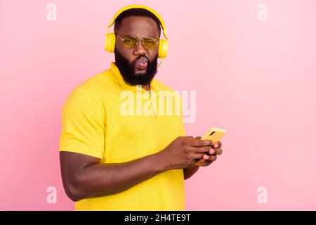 Photo of unhappy doubtful afro amnerican brunette haired stylish man hold phone listen music headset isolated on pink color background Stock Photo