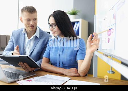 Young man and woman looking at screen of digital tablet and studying information on blackboard Stock Photo