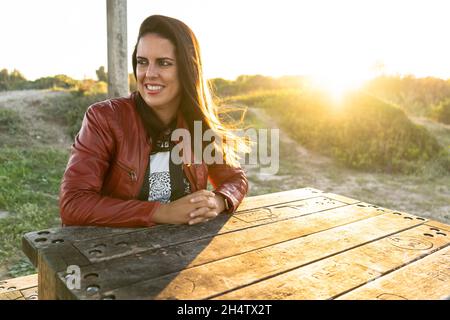 Young woman smiling sitting at a table in a park at sunset wearing a red jacket Stock Photo