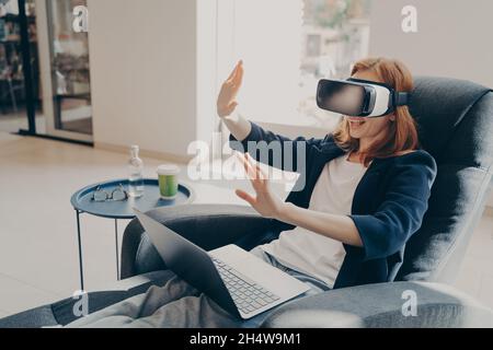 Young businesswoman wearing VR headset glasses and laptop sitting in cozy armchair in office lobby Stock Photo