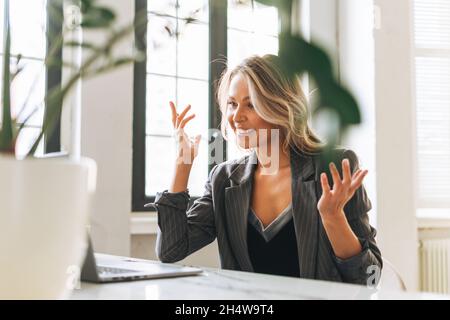 Young smiling blonde woman with long hair in stylish suit working at laptop in bright modern office, talking on video call Stock Photo