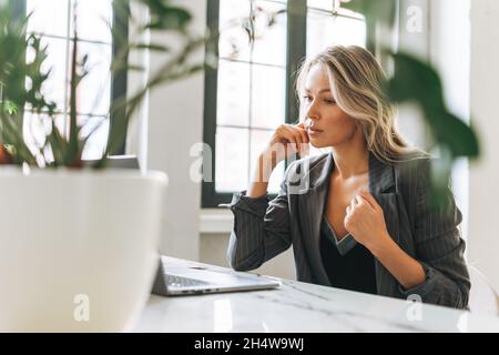 Young smiling blonde woman with long hair in stylish suit working at laptop in bright modern office, talking on video call Stock Photo