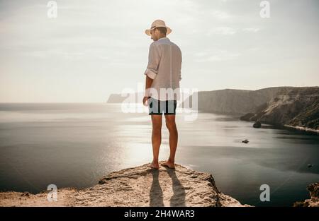 A digital nomad, a man in a hat, a businessman does yoga on the rocks by the sea at sunset time, does a business operation online from a distance Stock Photo