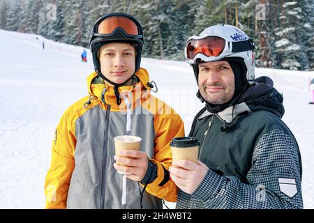 Two snowboarders of different ages are drinking hot coffee. Ski slope background. Father and son Caucasian ethnicity are engaged in winter extreme spo Stock Photo