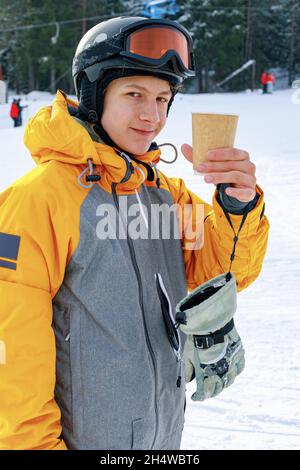 The snowboarder drinks hot coffee. A young guy of Caucasian ethnicity is holding a paper cup with a hot drink. Ski slope background. Portrait of an at Stock Photo