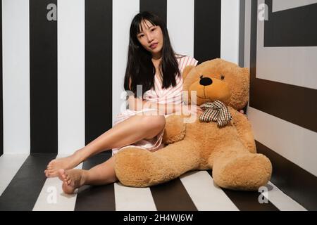 Young asian woman sitting with a teddy bear on the striped room floor. Stock Photo