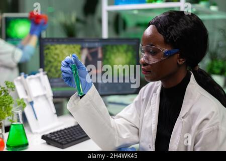 African american biologist researcher holding medical test tube with green solution working at biochemistry experiment in biochemistry hospital laboratory. Chemist analyzing geneticaly modified liquid Stock Photo