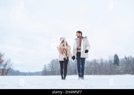 Young happy couple smile to each other and walk in winter park holding hands. Man and woman have fun together during winter holidays Stock Photo