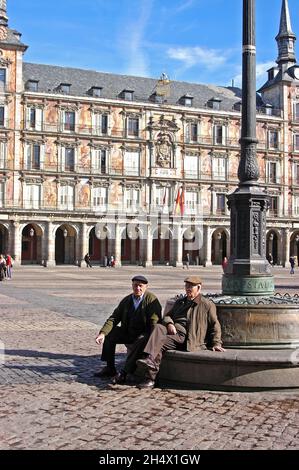 Two elderly Spanish men sitting on a bench with the facade of the Casa De La Panaderia to the rear, Plaza Mayor, Madrid, Spain. Stock Photo