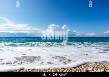 Scenic white foam on pebble beach with blue stormy waves and epic cloudscape on coast of Lefkada island in Greece. Summer nature travel to Ionian Sea Stock Photo