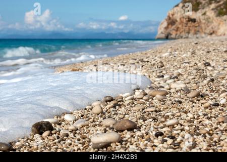 White foam on wet shiny pebble stones close-up with azure sea, stormy waves, epic clouds and rocky cliff. Mylos beach on coast of Lefkada island in Gr Stock Photo