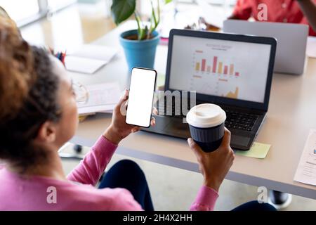 Businesswoman holding coffee cup while looking at smartphone with copy space in office Stock Photo