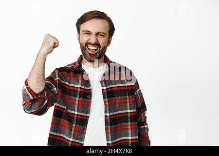 Portrait of enthusiastic bearded man raising clench fist and rooting for team, enjoying watching game, standing over white background Stock Photo