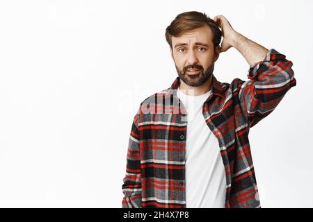 Confused caucasian bearded man scratch head, looking clueless, dont understand smth, standing in checked shirt over white background Stock Photo