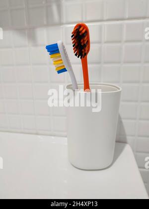 Two colored toothbrushes in white plastic glass on the shelf. Stock Photo