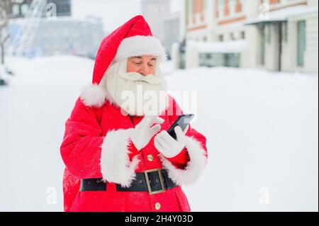 Surprised santa claus is using mobile outside. Santa Claus stands in the background of buildings in winter and calls on a smartphone. Snow in December Stock Photo