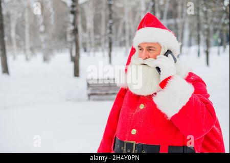 Surprised santa claus is using mobile outside. Santa Claus stands in the background of buildings in winter and calls on a smartphone. Snow in December Stock Photo
