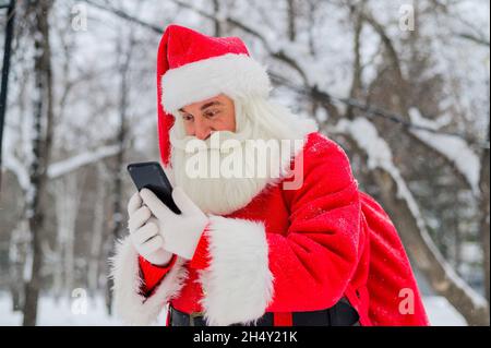 Surprised santa claus is using mobile outside. Santa Claus stands in the background of buildings in winter and calls on a smartphone. Snow in December Stock Photo