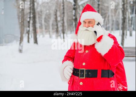 Surprised santa claus is using mobile outside. Santa Claus stands in the background of buildings in winter and calls on a smartphone. Snow in December Stock Photo