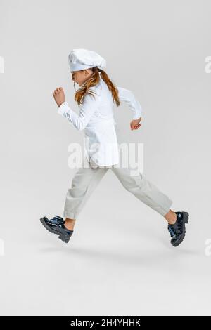 Little Caucasian girl in white cook uniform and huge chef's hat running isolated on white studio background. Stock Photo