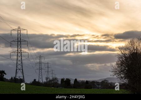 High-voltage, overhead power lines and pylons carrying electricity energy supplies through UK countryside setting with winter sunset sky. Stock Photo