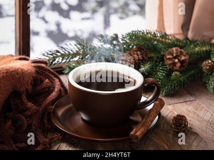 Steaming hot cup of coffee on a rustic wooden table by a window with winter outdoors background Stock Photo