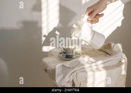Pouring coffee from Italian coffee maker with wooden handle into grey espresso craft cup on solid wood stump decorated with linen tea towel in creamy