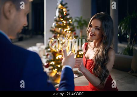 Stunning woman in red dress and handsome man in stylish suit toasting with champagne while celebrating christmas eve at home.Concept of winter holidays and relationship. Stock Photo