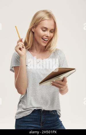 Young joyful european girl watch something in notebook paper and have new idea. Beautiful blonde female teenager with blue eyes wear white t-shirt and Stock Photo