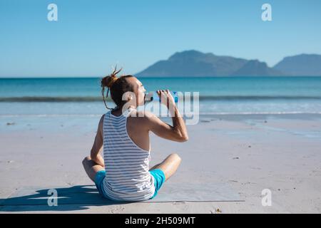 Rear view of man drinking water from bottle while doing yoga at beach on sunny day Stock Photo