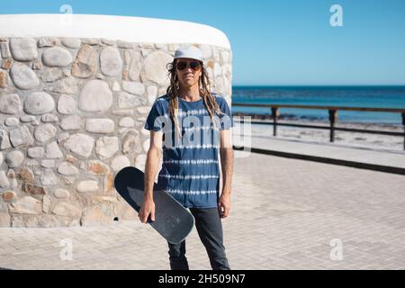 Portrait of male hipster holding skateboard against wall at promenade on sunny day Stock Photo