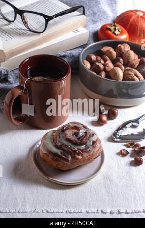 Cosy autumn scene with a freshly baked cinnamon roll, hot tea, a variety of nuts, books, a pumpkin and a persimmon. Stock Photo