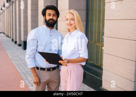 Portrait of smiling male and female colleagues holding clipboard standing posing in city street by office building, looking at camera. Stock Photo