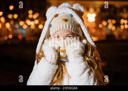 Close-up portrait young girl in knitted hat holds her hands in mittens near her face, covers her face with scarf from cold outside. Girl in evening on christmas street in light of lights and garlands Stock Photo