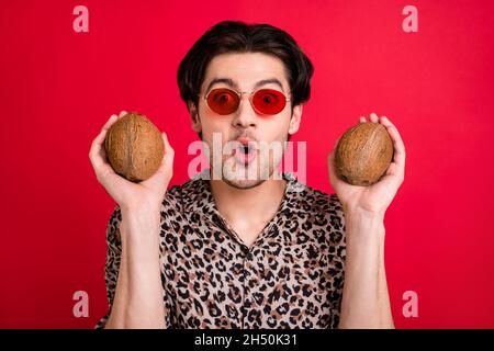 Portrait of young funky good mood amazed man in sunglass hold two coconuts tropical fruit isolated on red color background Stock Photo