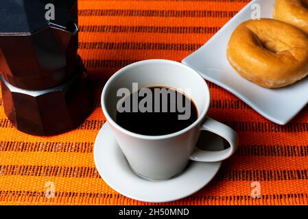 Cup of coffee next to a coffee pot with some glazed donuts on a white plate. Stock Photo