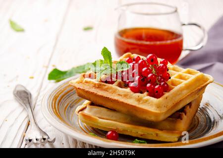 Belgian waffles served with red currants and mint leaf on white wooden kitchen table with syrup aside Stock Photo