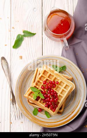 Flat lay view at Belgian waffles served with red currants and mint leaf on white wooden kitchen table with syrup aside Stock Photo