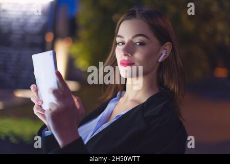 Young businesswoman with hands free device using tablet at night in city, looking into camera Stock Photo