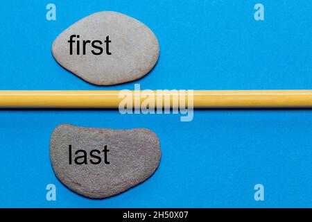 The words first and last are written on natural smooth stones separated by a yellow pencil. The background is isolated in blue and has a lot of space Stock Photo