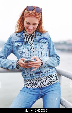 Happy young redhead Caucasian woman messaging on smartphone at riverbank Stock Photo