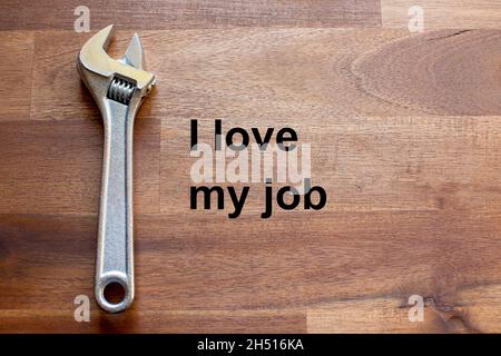 a flexible chromed adjustable wrench lies on a dark wooden table. The solgan I love my job is written on the table in black letters Stock Photo