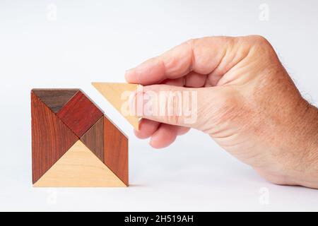 The missing building block is inserted into a tangram made of fine wood by a senior's hand Stock Photo