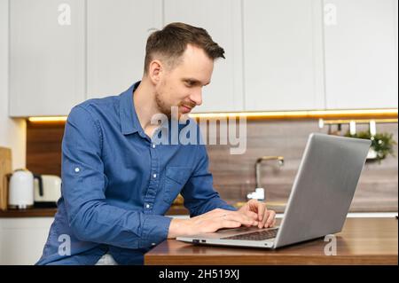 Concentrated serious male entrepreneur using laptop sitting in the kitchen at home, confident good looking man sending emails, typing on the keyboard, working remotely Stock Photo